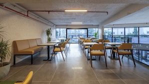 Amplia zona de estar interior con mesas y sillas, ventanas grandes que ofrecen vistas de la ciudad y un techo alto con tuberías a la vista.
