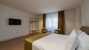 Une chambre d’hôtel comprenant un lit, un téléviseur fixé au mur, un coin salon avec des fauteuils et une fenêtre avec des rideaux.