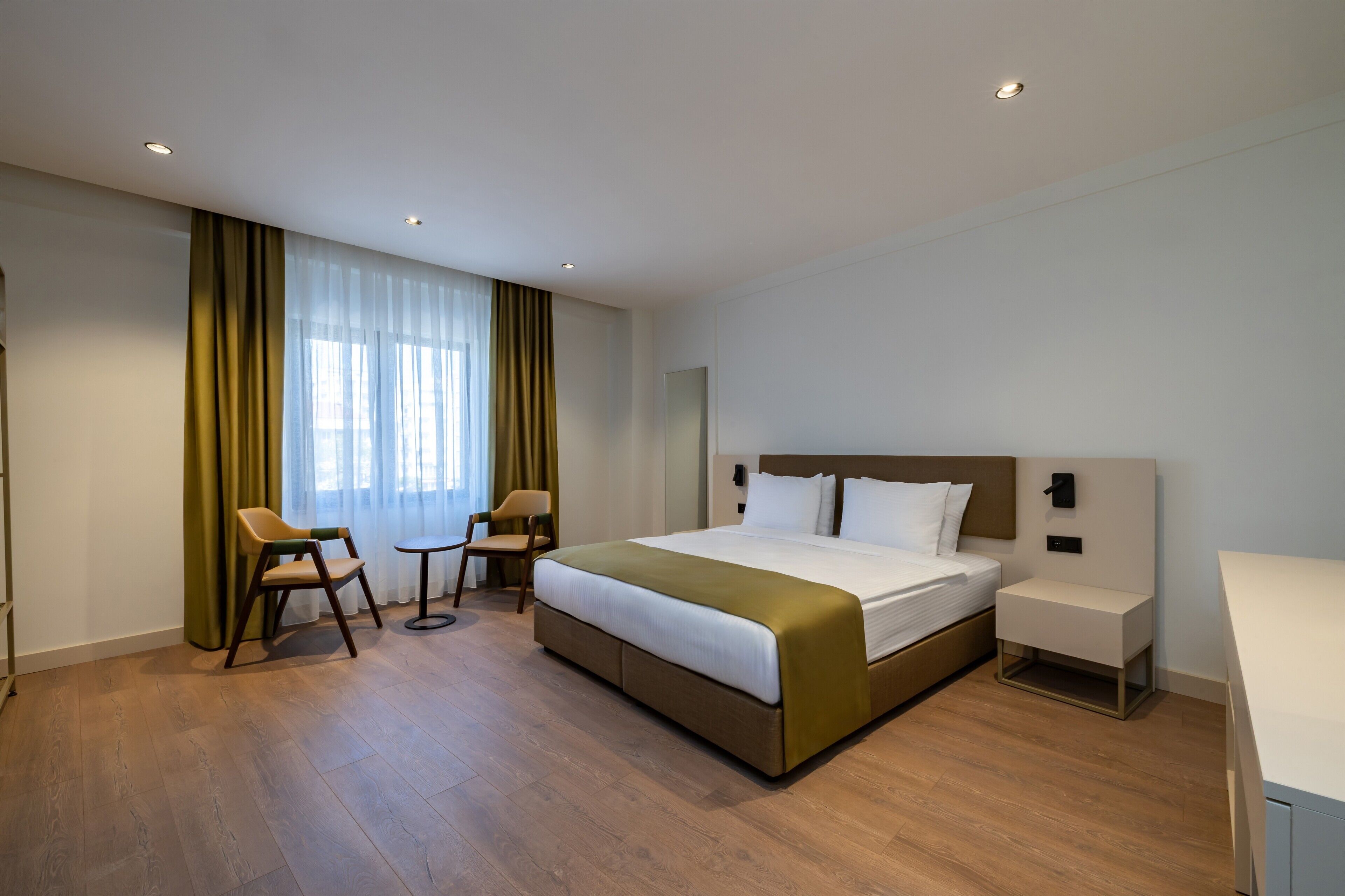 Quarto de hotel com cama, escrivaninha, duas cadeiras, janela com cortinas e piso de madeira.