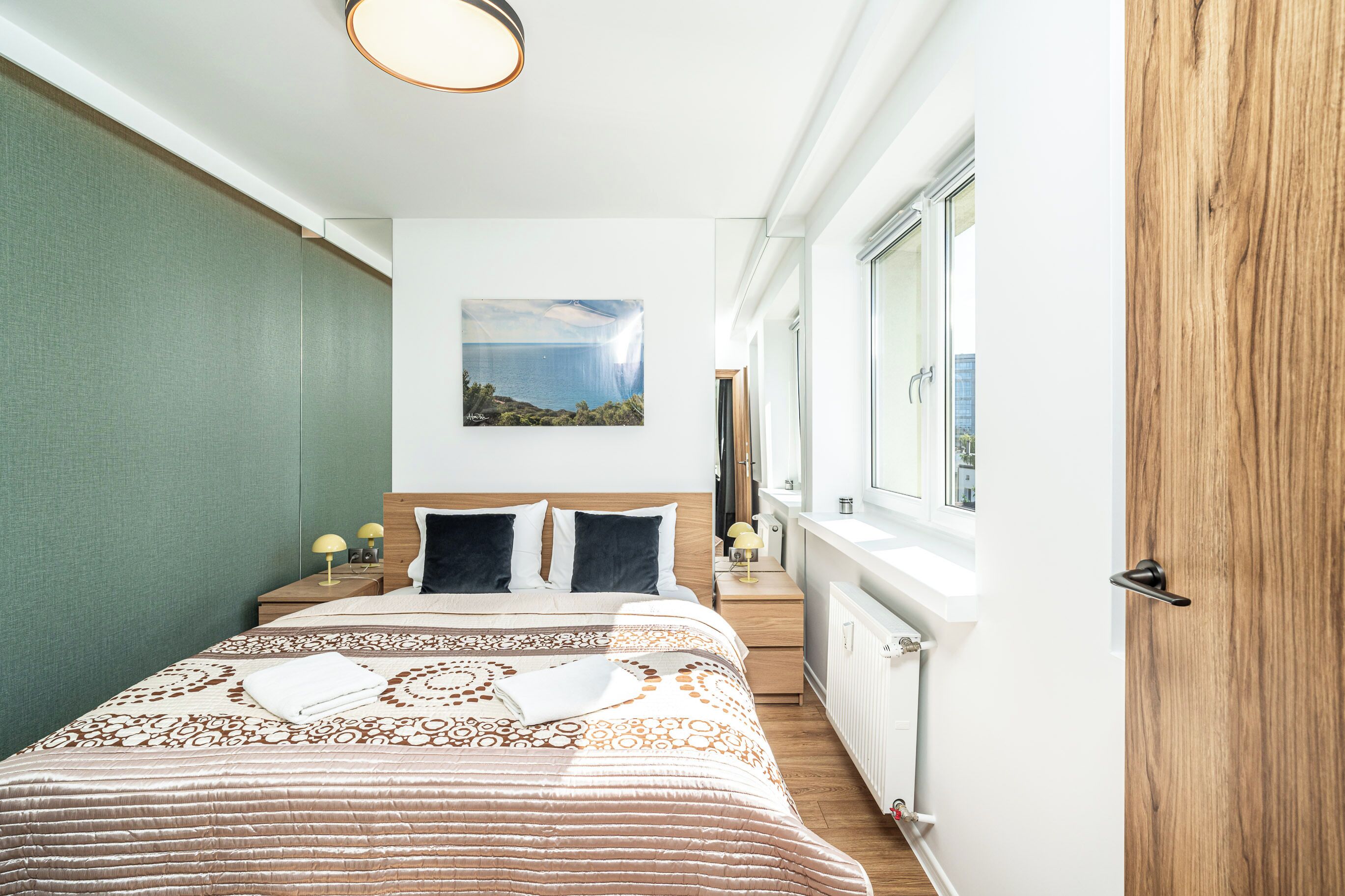 Une chambre d’hôtel étroite, avec un lit, des lampes de chevet, un tableau et une fenêtre donnant sur un paysage.