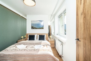 A narrow hotel room with a bed, bedside lamps, a painting, and a window with a view. - RentPlanet - Apartament Cicha (Katowice)