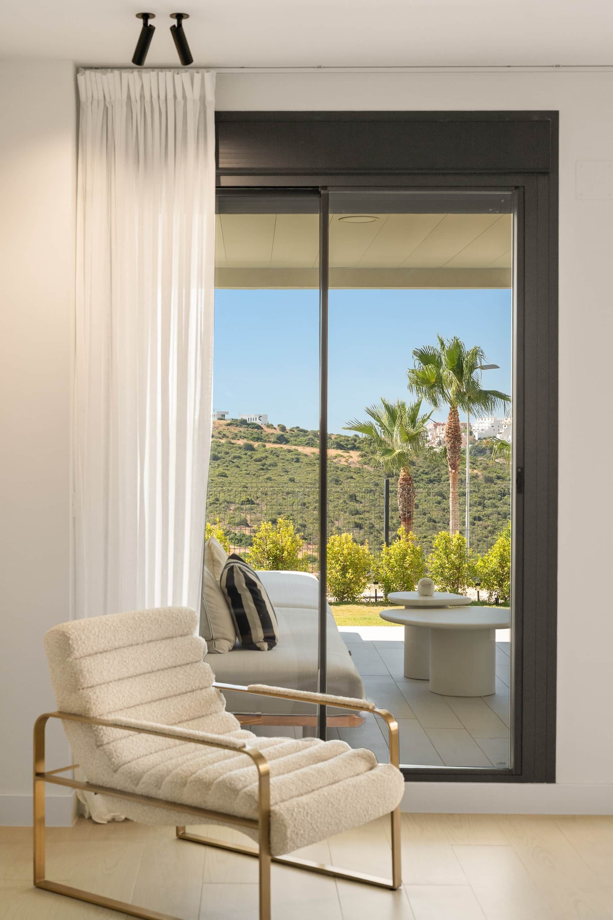 A modern room with a white armchair, a large sliding glass door leading to an outdoor area with a view of a hilly landscape and palm trees.