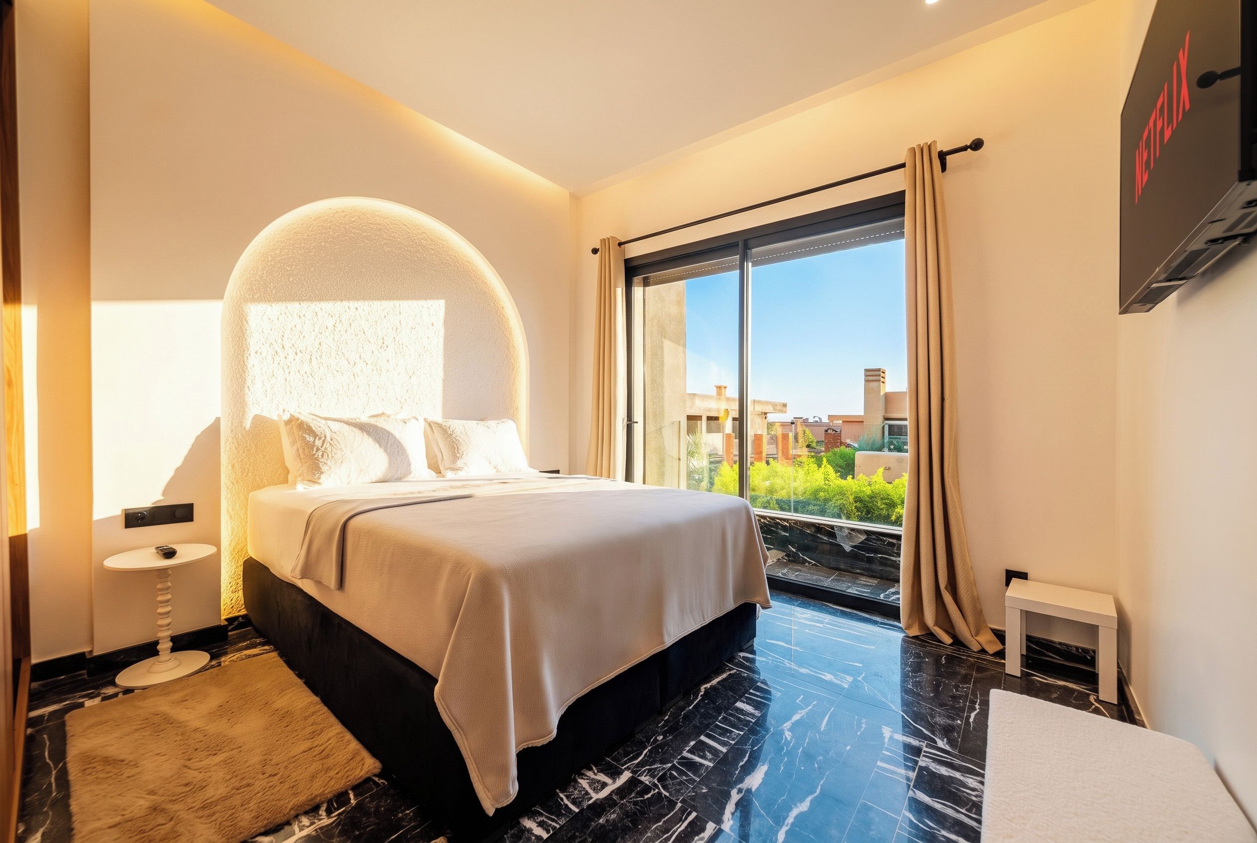 A modern hotel room with a large bed, a round mirror above the bed, a large window with curtains, and a flat-screen TV mounted on the wall.