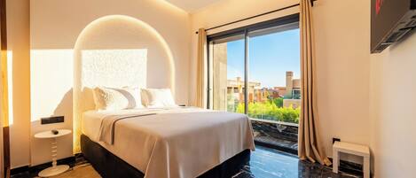 A modern hotel room with a large bed, a round mirror above the bed, a large window with curtains, and a flat-screen TV mounted on the wall.