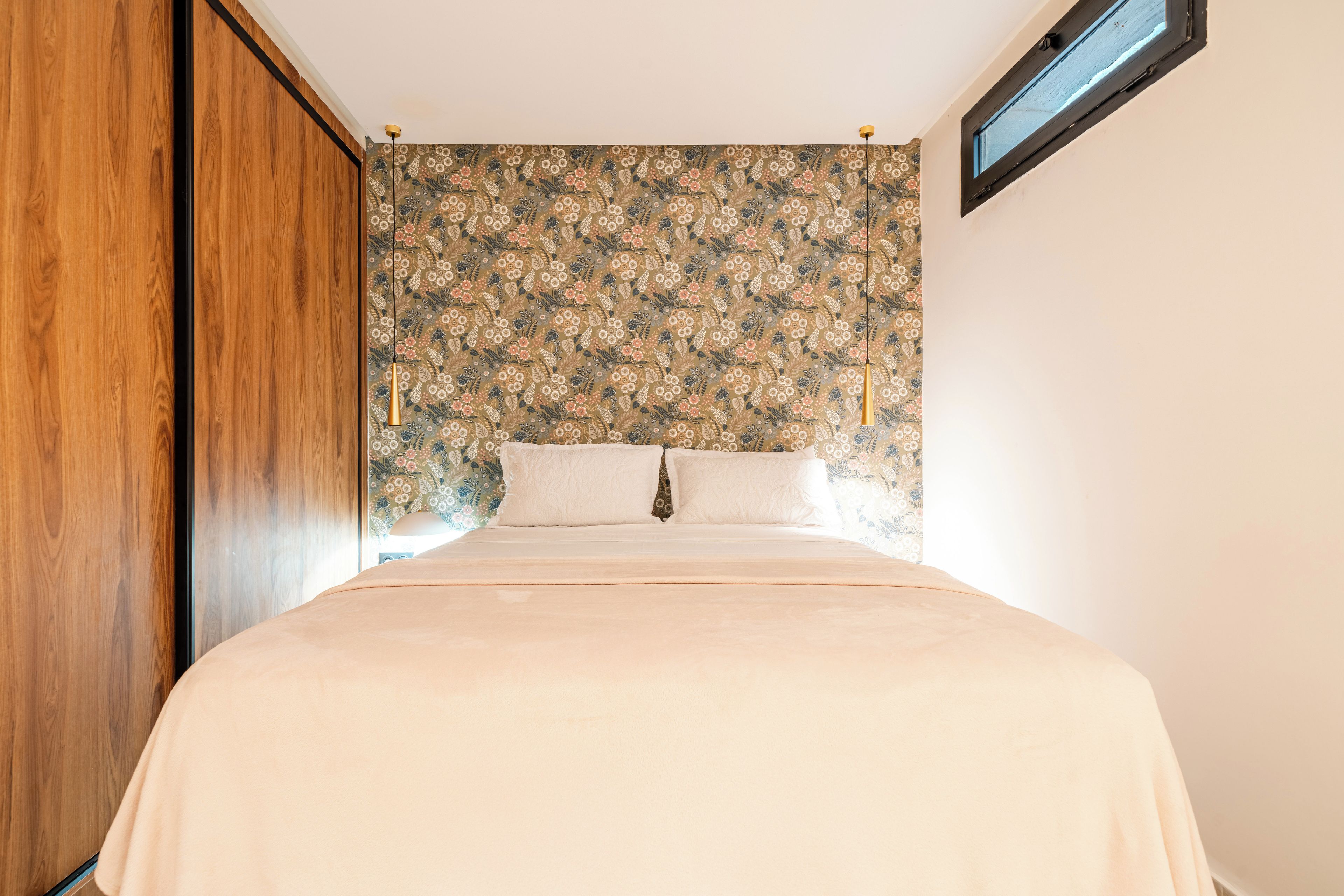 A bedroom with a bed, wooden wardrobe, floral wallpaper, and a window.