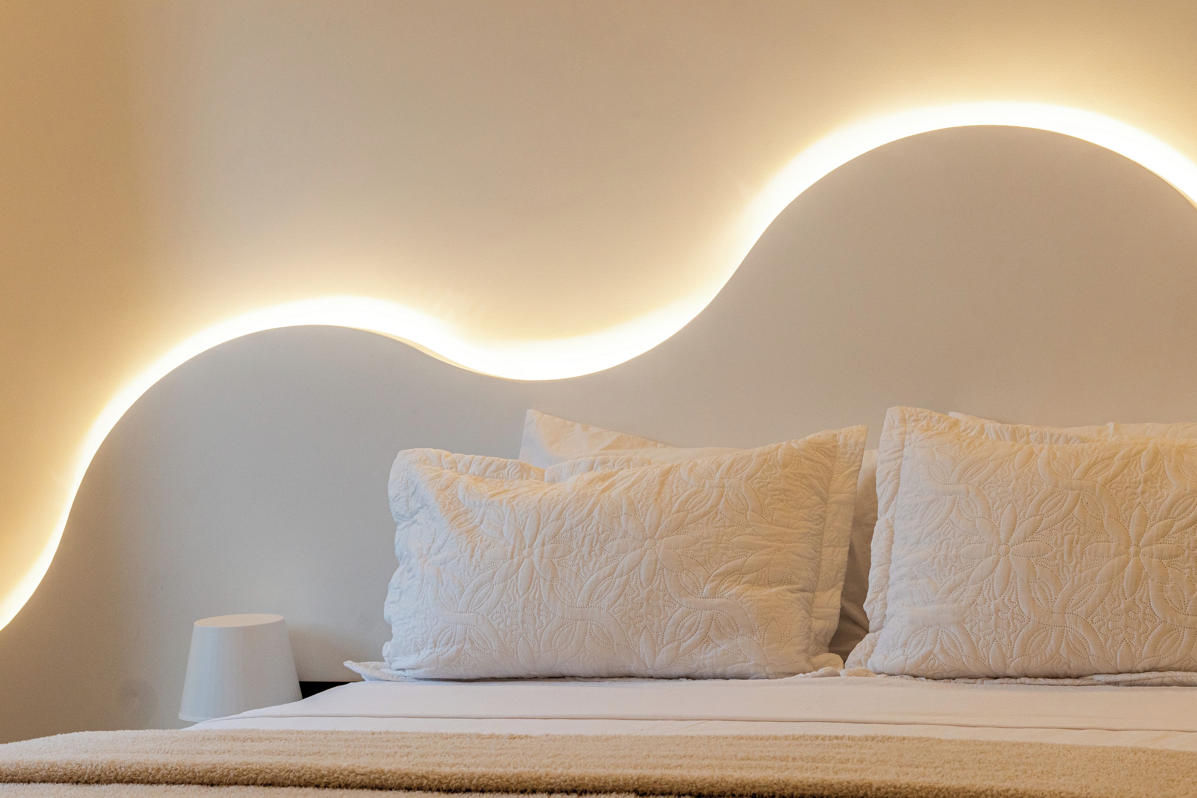 Ein Bett mit weißer Bettwäsche und Kissen, eine Nachttischlampe und eine Wand mit einer Leuchte.