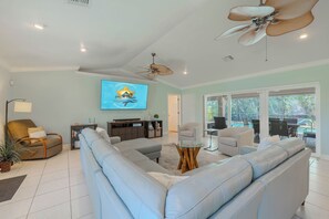 A spacious living room with a large L-shaped sofa, a television mounted on the wall, a ceiling fan, and a view of an outdoor pool area through large windows. - Sleeps 8 - 2 bath- Pool and Hot Tub - Pet Friendly (Cape Coral)