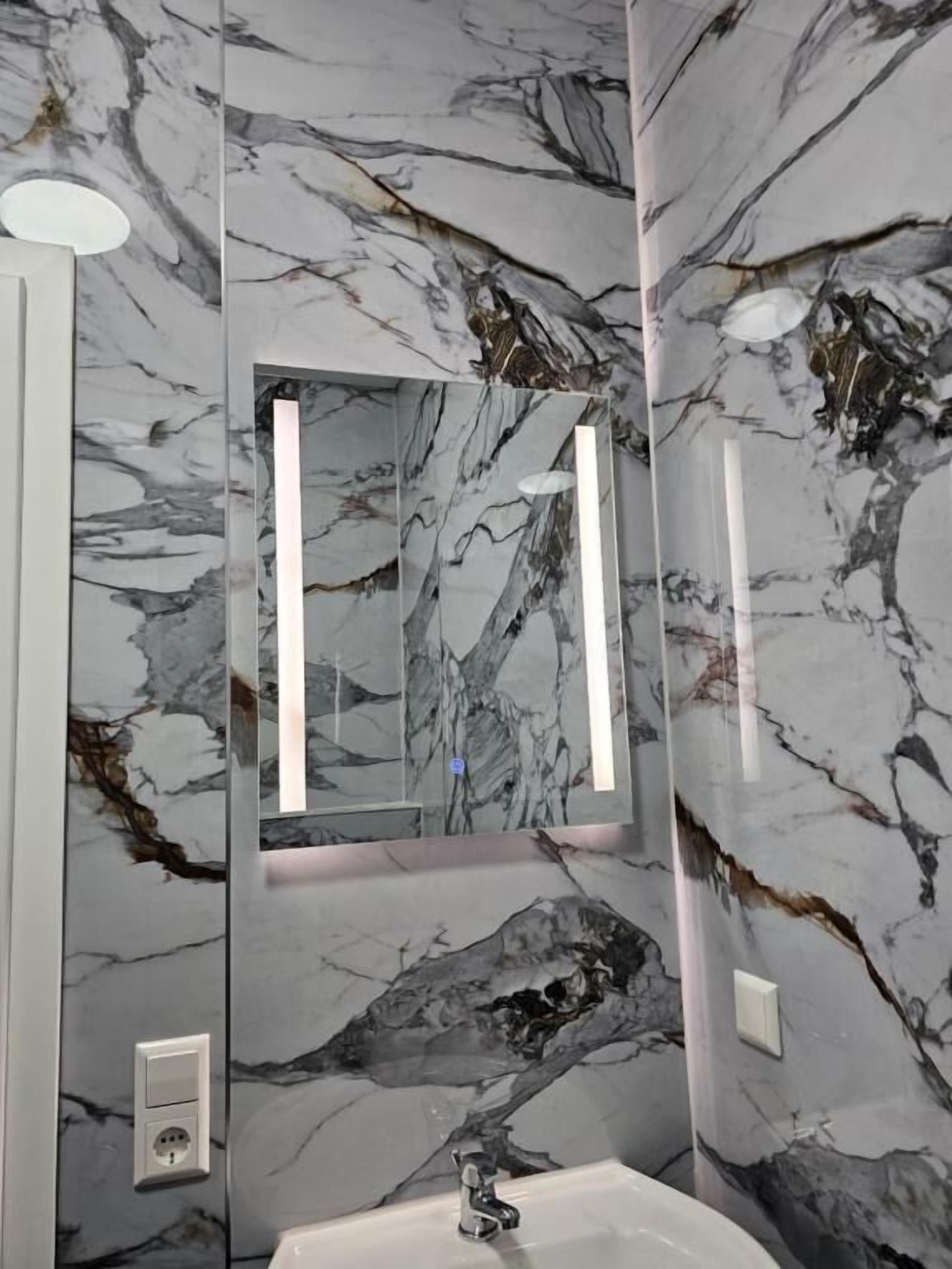 Un baño con paredes de mármol, un lavamanos blanco y un espejo con iluminación vertical.