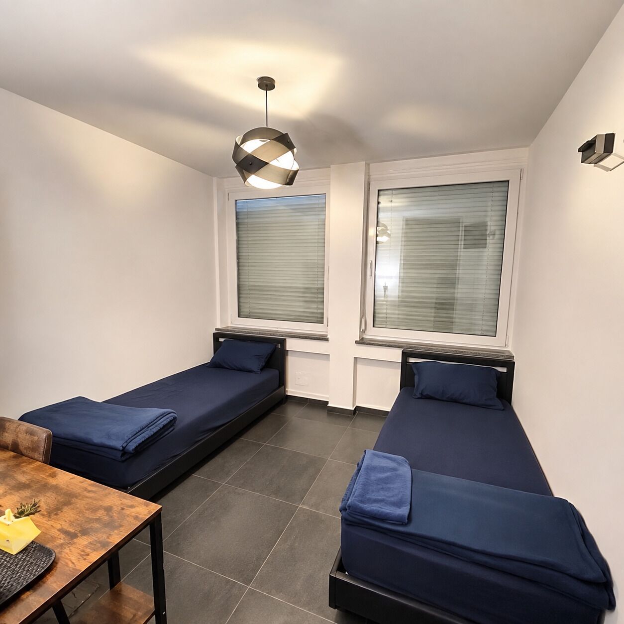 Ett litet, modernt rum med två enkelsängar, ett bord och en hängande lampa.