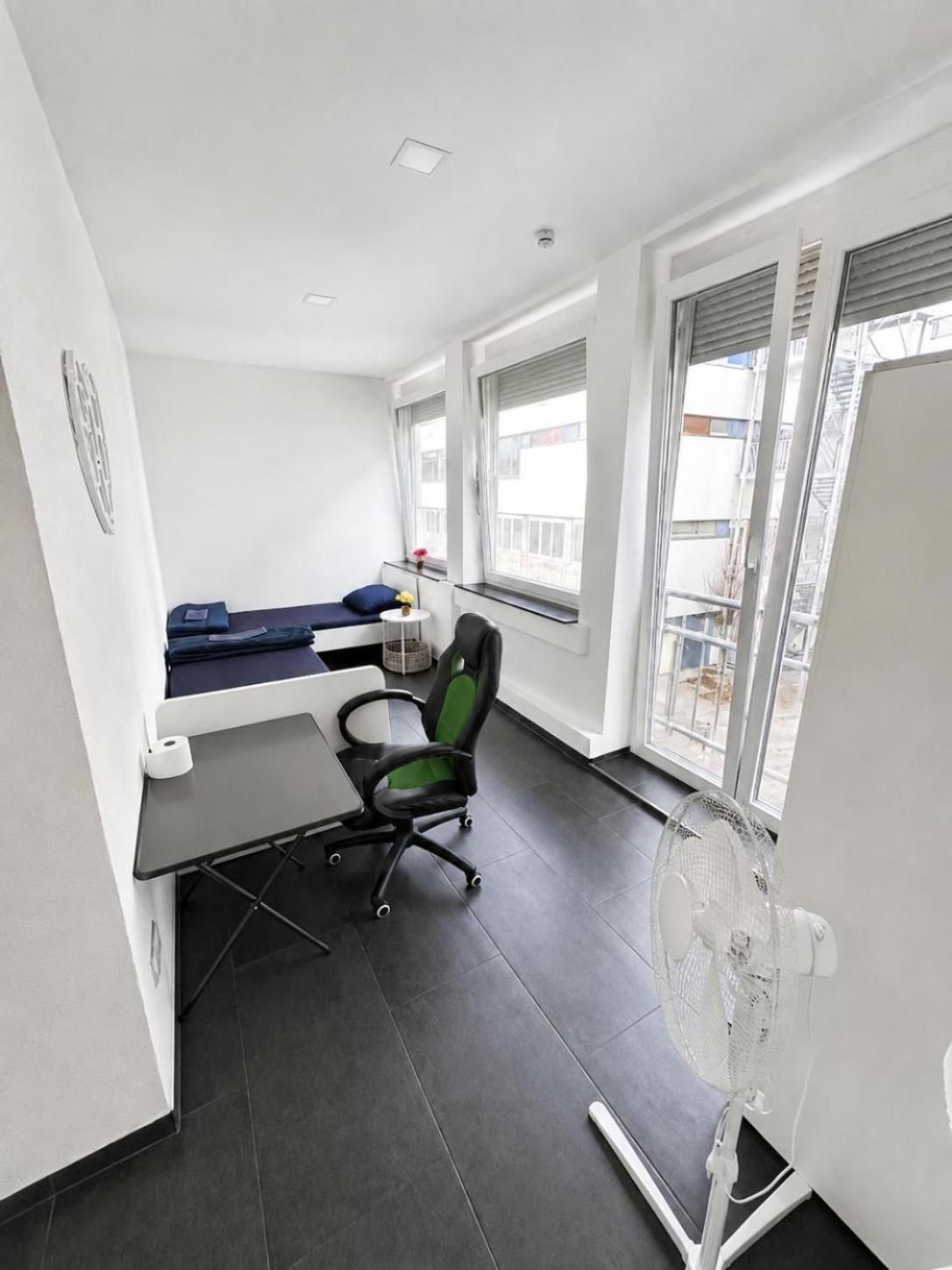 Un espacio de oficina moderno con un escritorio, una silla y una pequeña mesa con un rollo de papel. Hay una ventana con persianas, un balcón visible a través de la ventana y un ventilador de pie.