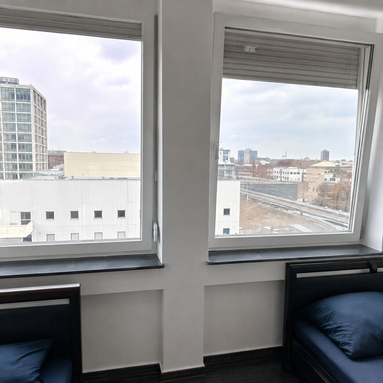 Ein Zimmer mit zwei Betten, einem großen Fenster mit Blick auf Gebäude und Gleise sowie einer weißen Wand.