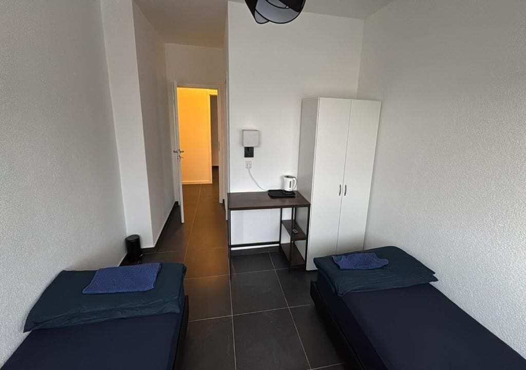 Et lille, kompakt værelse med to senge, et skrivebord med stol, et garderobeskab og en lampe på væggen.