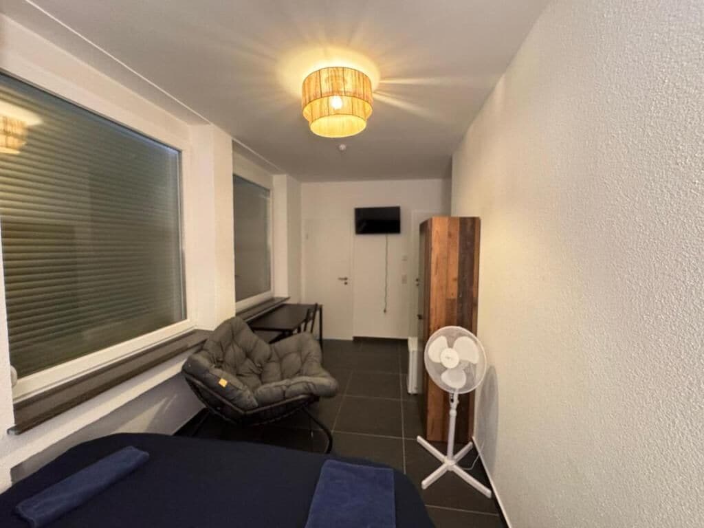 Ein Zimmer mit einem Bett, einem Sessel, einem Standventilator, einem an der Wand befestigten Fernseher und einem Fenster mit Jalousien.