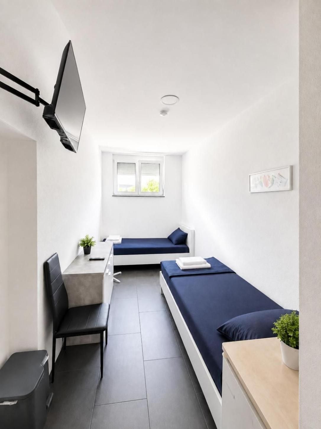 Il s’agit d’une pièce de taille modeste, comprenant un lit, un bureau et une chaise. On y trouve une fenêtre avec des stores, un téléviseur fixé au mur et une petite plante posée sur le bureau.