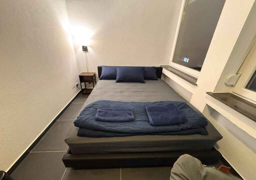 Una habitación pequeña con cama, mesita de noche y ventana.