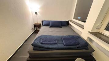 Una habitación pequeña con cama, mesita de noche y ventana.