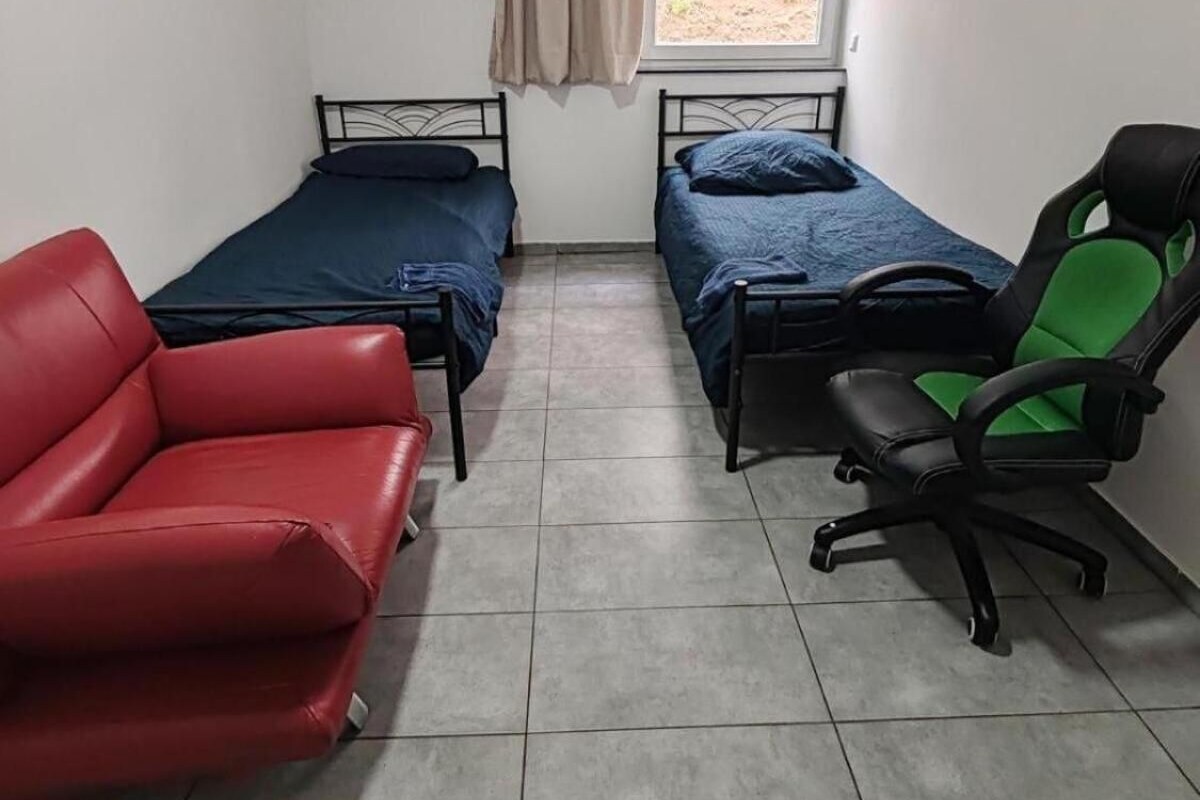 Un alojamiento pequeño, una habitación individual con dos camas, una silla roja, una silla de oficina verde y negra, y una ventana con cortina.