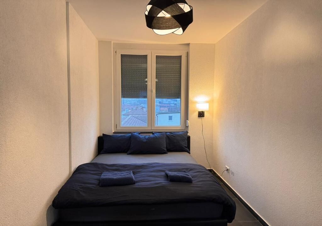 A narrow hotel room with a bed, a window with blinds, a wall-mounted lamp, and a view of the cityscape.