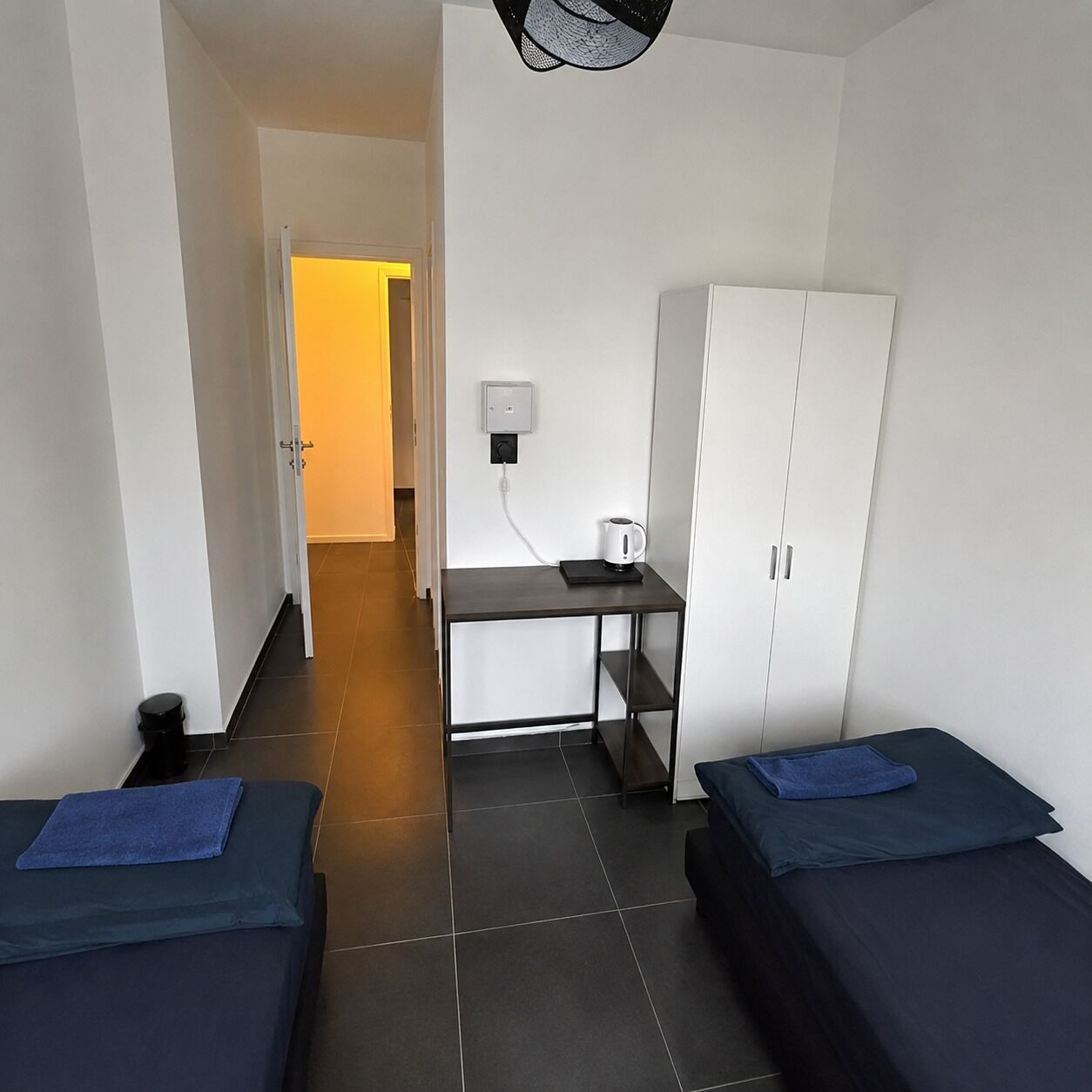 A hotel room with two beds, a desk with a kettle, a wardrobe, and a power outlet on the wall.