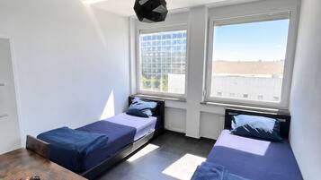 Ein kleiner, moderner Raum mit zwei Betten, einem Tisch und einem Fenster mit Blick auf ein Gebäude.