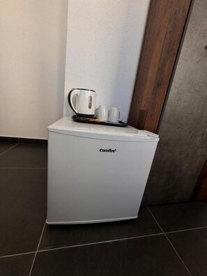 A compact coffee machine with a white exterior, placed on a tiled floor against a wall with a wooden panel.