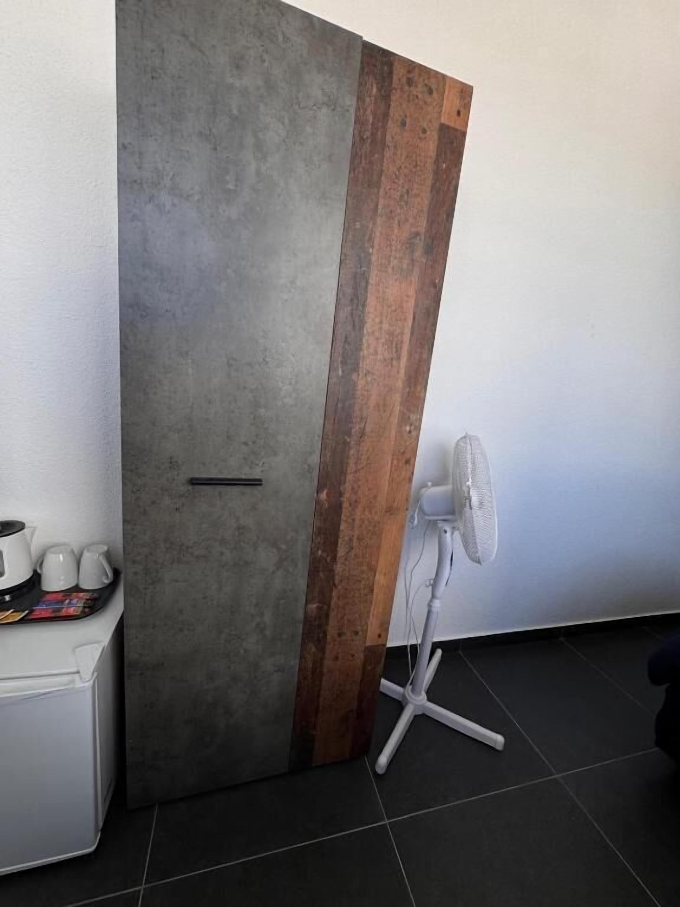 A room with a grey concrete wall, a wooden accent panel, a white fan, and a small white cabinet with cups.