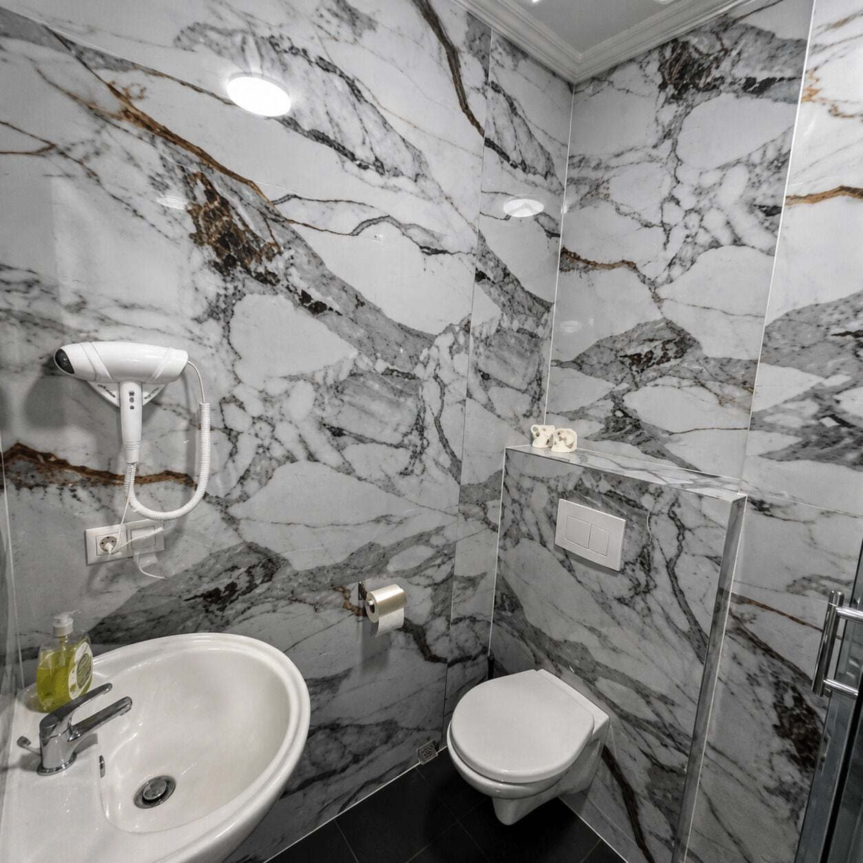 A bathroom with a marble wall, a sink, a toilet, and a hairdryer mounted on the wall.