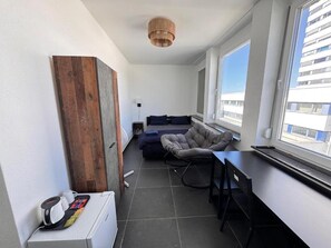 A modern living space with a sofa, a chair, a desk, and a large window offering a view of the city.
