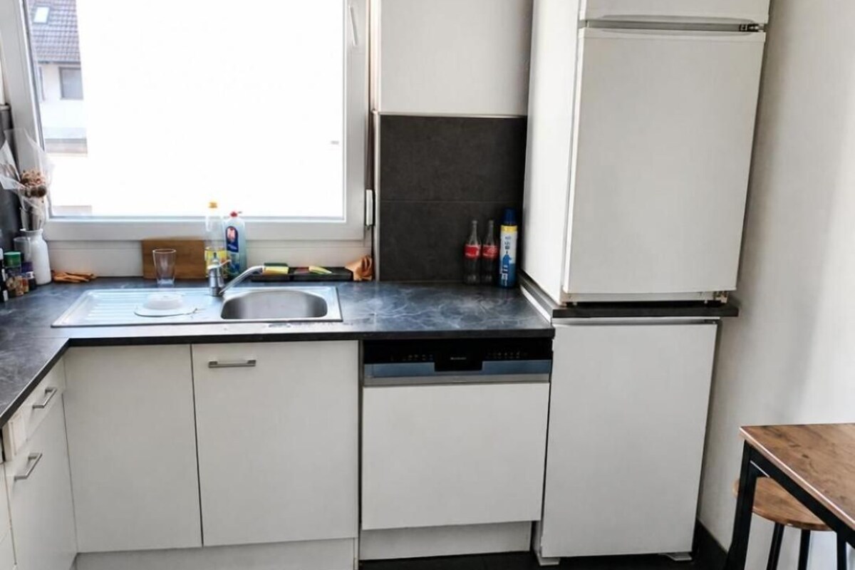 Una cocina compacta con lavavajillas integrado, un refrigerador, un ventanal que proporciona luz natural y un pequeño comedor con un taburete.