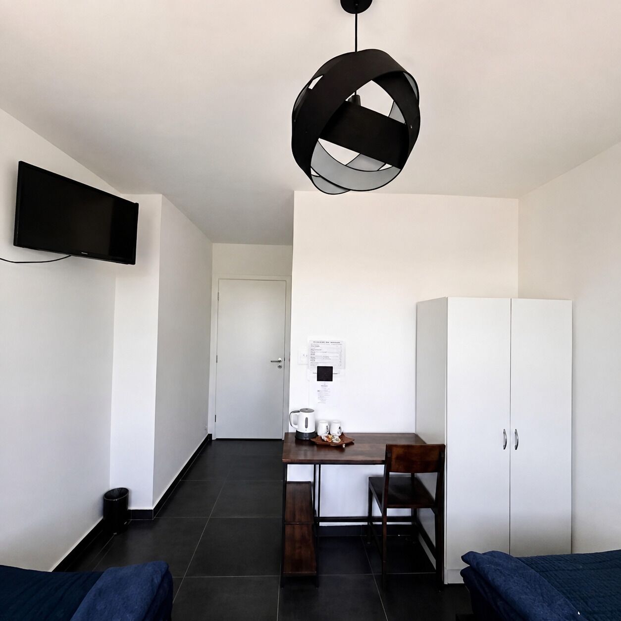 Quarto com uma cama, uma escrivaninha com cadeira, um guarda-roupa, uma televisão fixada na parede e um lustre.