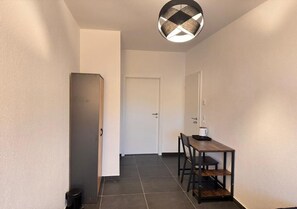 A hallway with a door, a trash bin, a table with chairs, and a hanging light fixture.