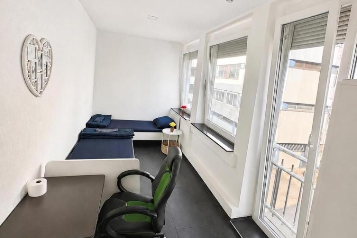 Una habitación pequeña y moderna con una cama, un escritorio y una silla. Una ventana ofrece vistas al exterior.