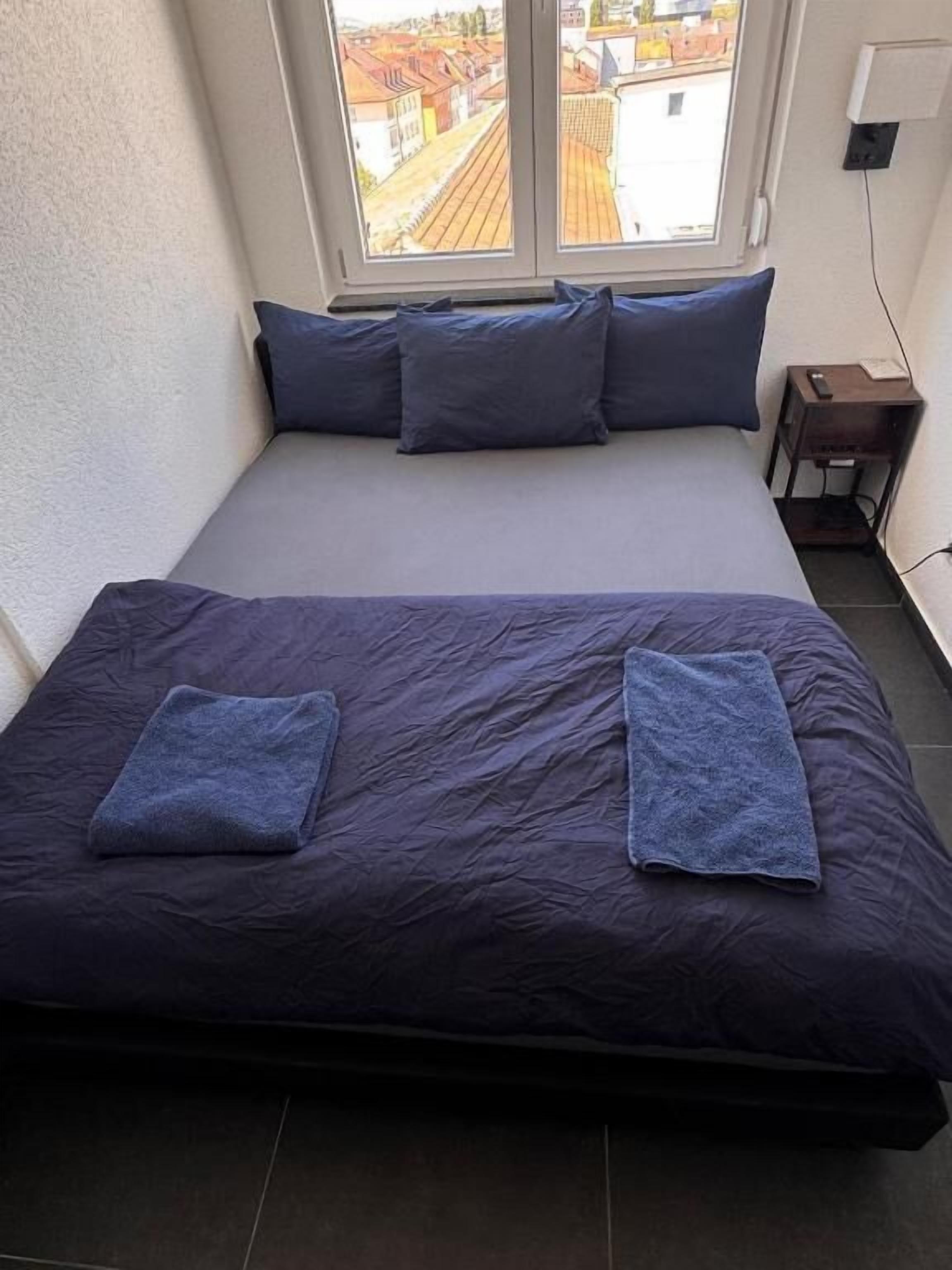 A bed with a dark-colored mattress and pillows, a small bedside table, and a window with a view of buildings.