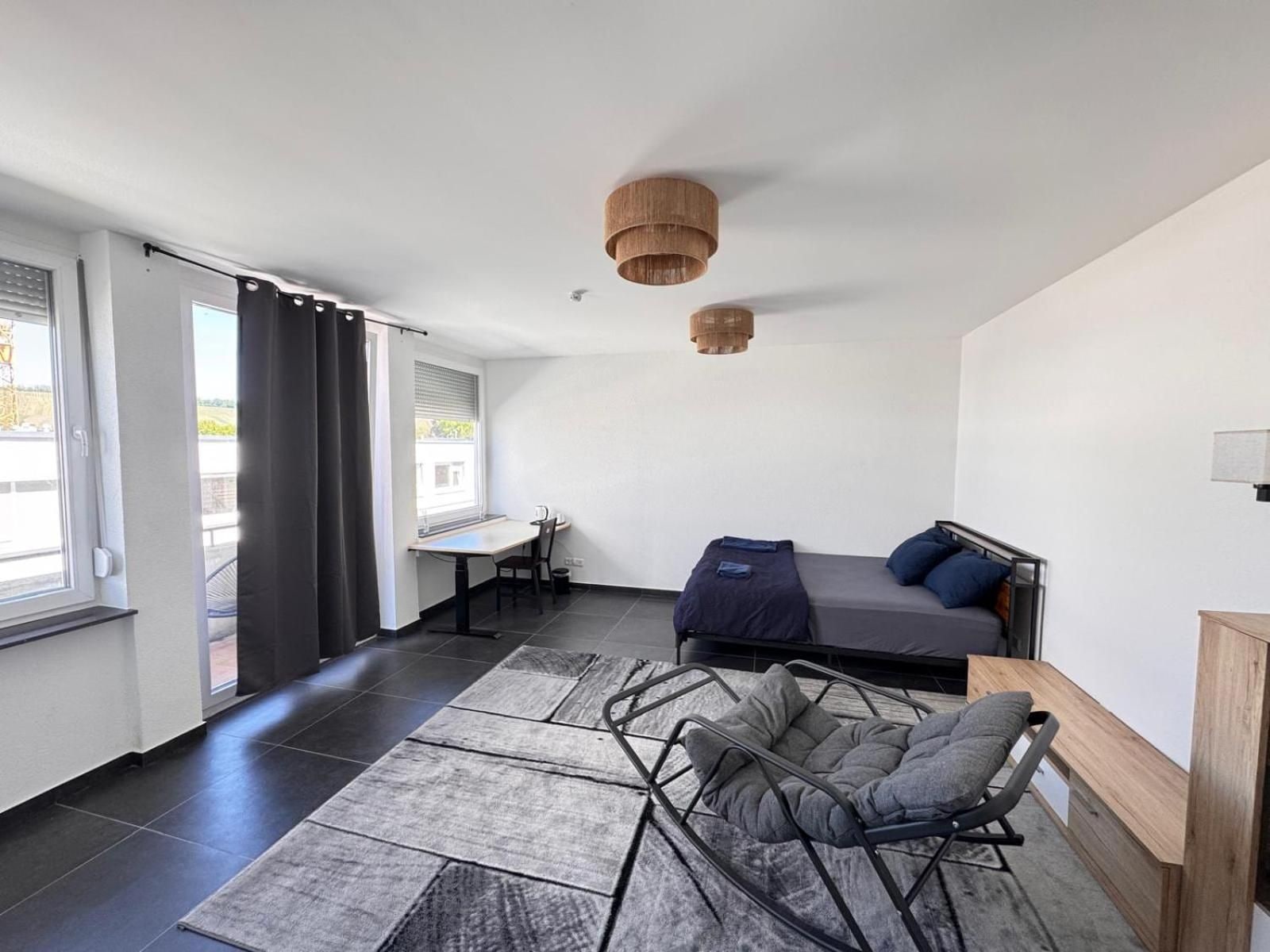 Ein modernes Zimmer mit einem Bett, einem Schreibtisch und einem Stuhl. Es gibt ein Fenster mit Vorhängen, eine Pendelleuchte und einen Teppich auf dem Boden.