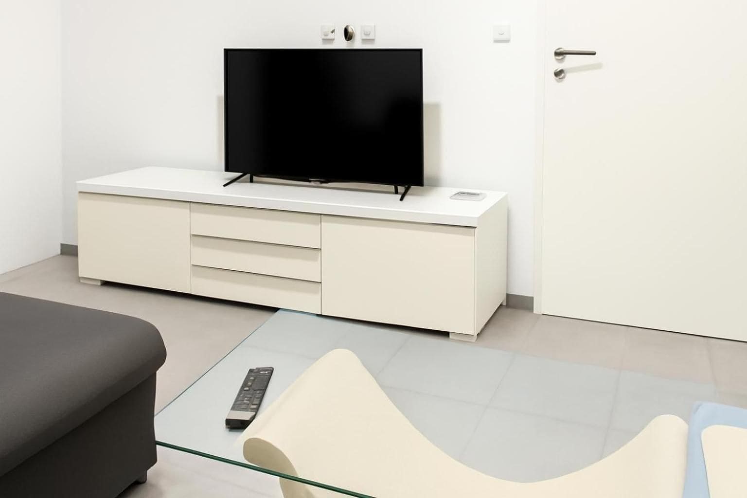 A modern room with a flat-screen TV on a white cabinet, a glass coffee table with a remote, and a door with a handle.