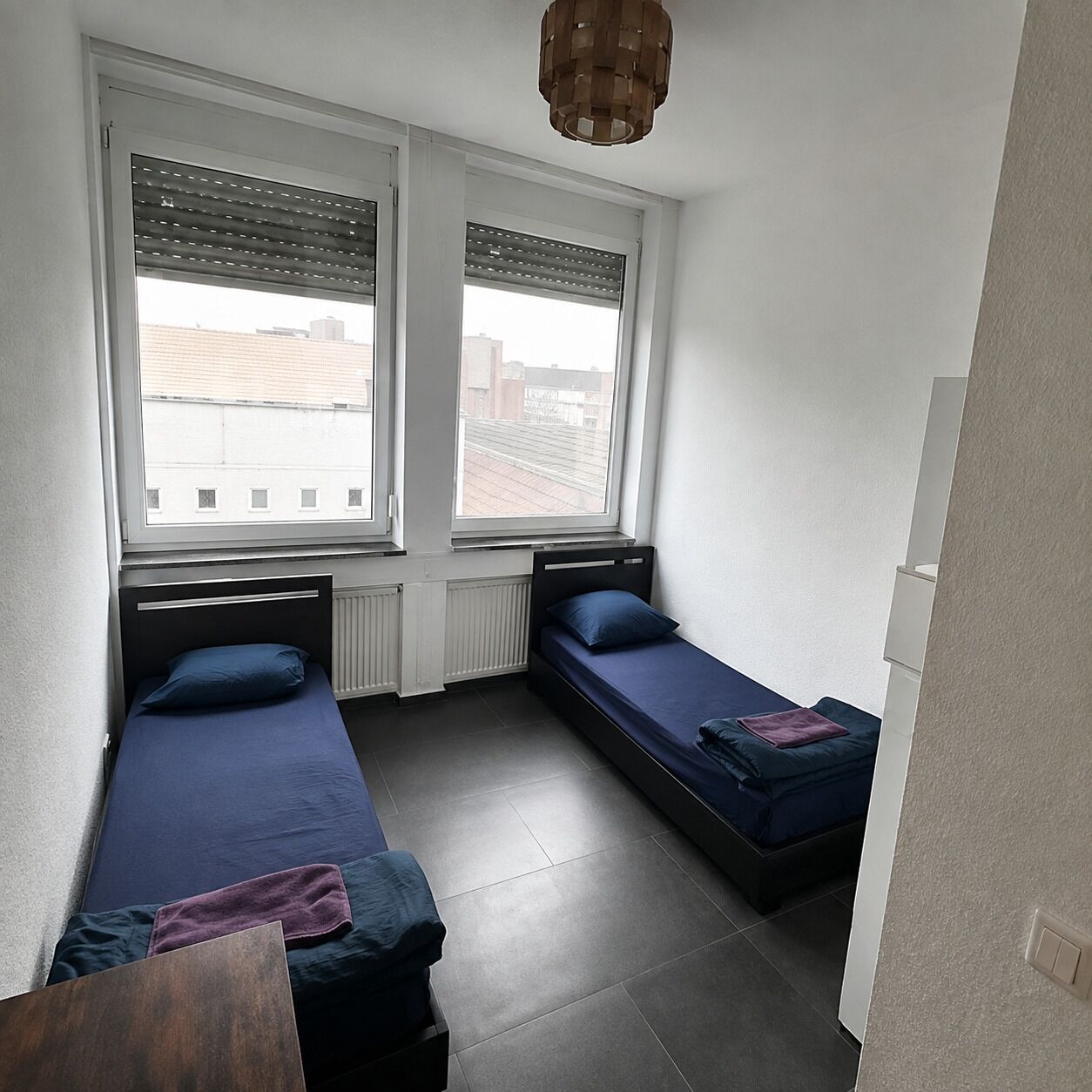 A small, compact room with two single beds, a radiator, and a window with blinds.