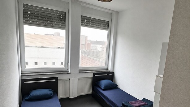 Una habitación pequeña y funcional con dos camas individuales, un radiador y una ventana con persianas.