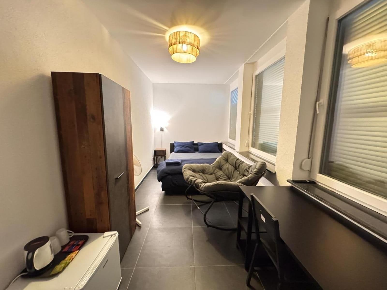 Une petite pièce comprenant un lit, une chaise, une table et une fenêtre avec des stores.