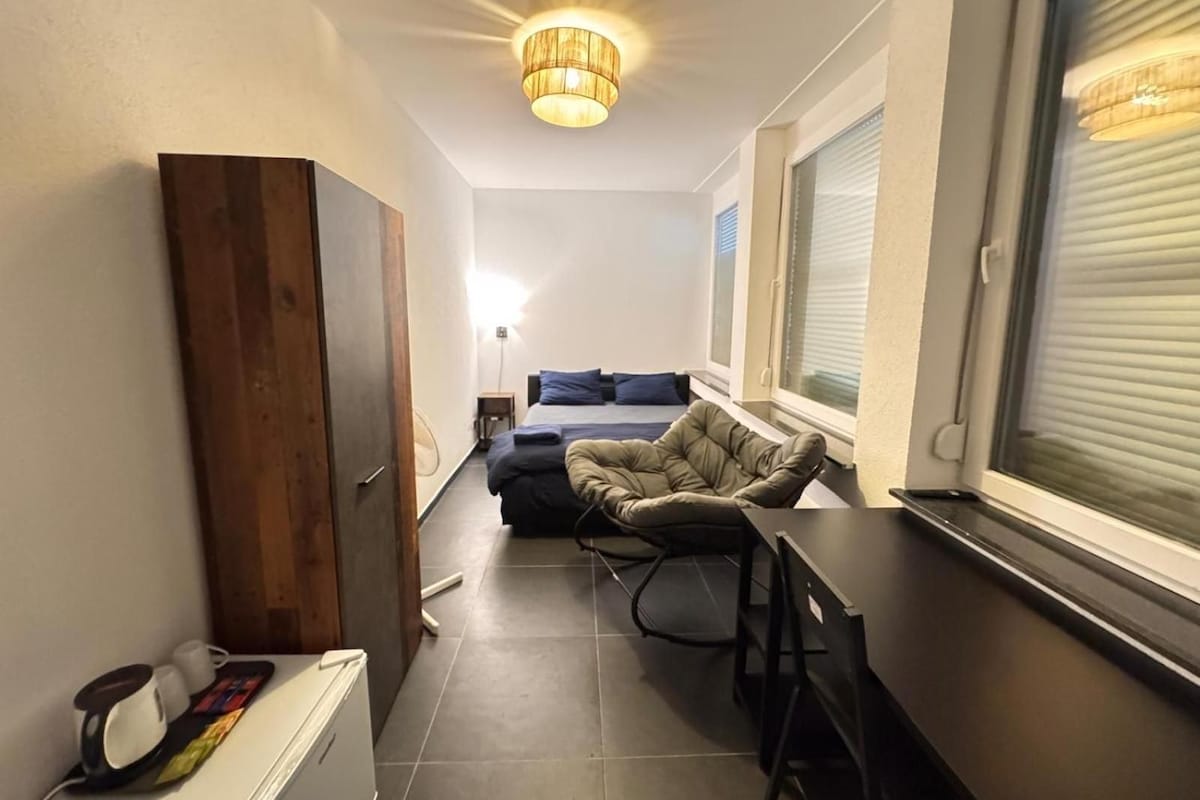 Una habitación pequeña con una cama, una silla, una mesa y un ventanal con persianas.