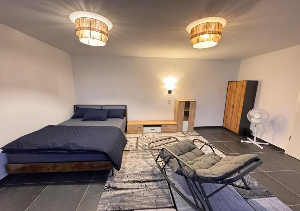 Ein Schlafzimmer mit Bett, Sessel, Nachttisch, Kleiderschrank und einer Wandleuchte.