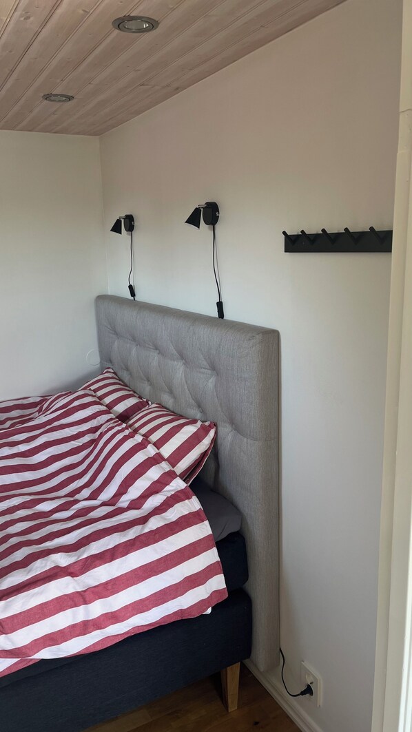 A bedroom with a bed, a headboard, a wall-mounted lamp, and a power outlet.