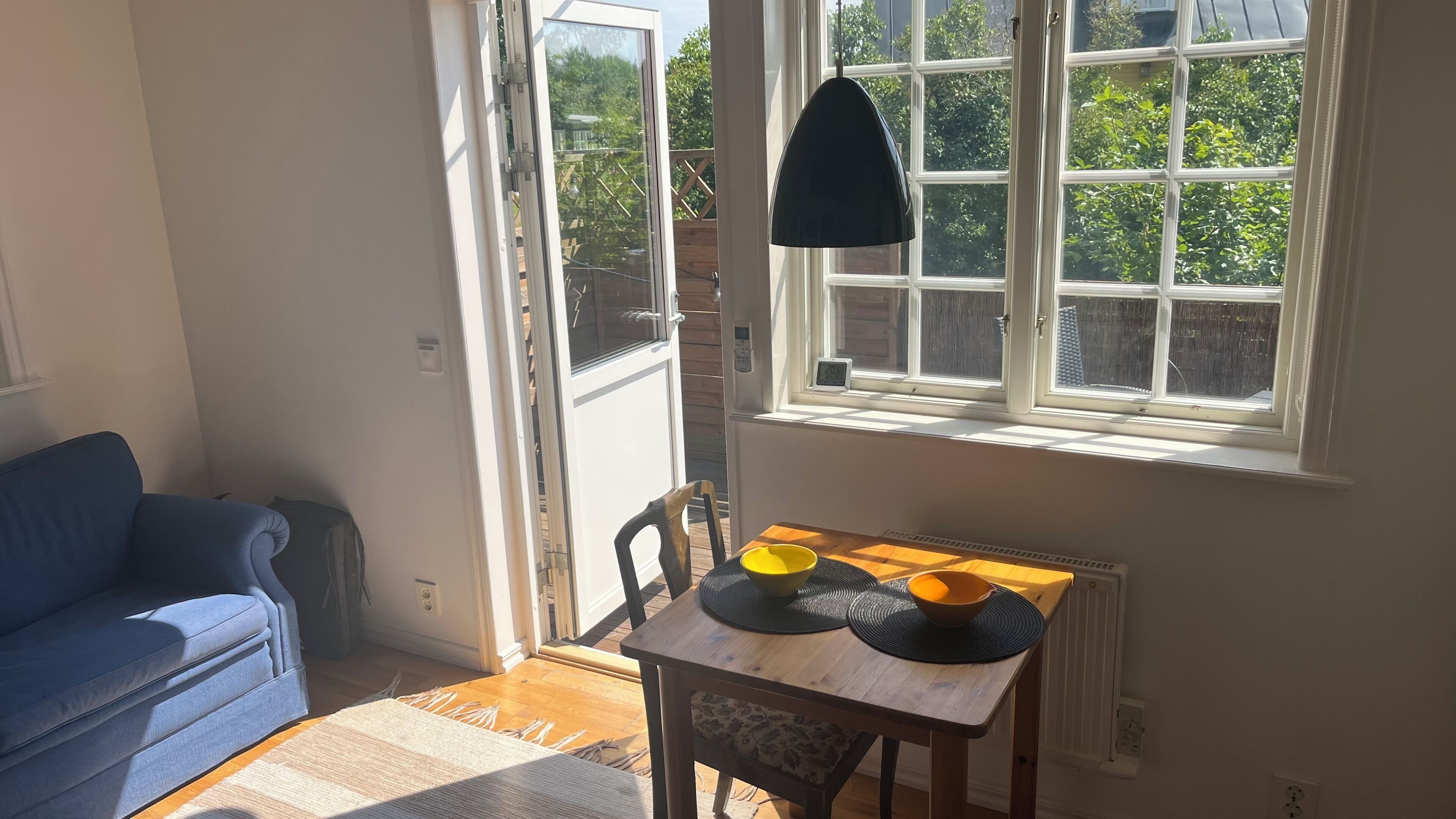 Una habitación con un sofá azul, una mesa de madera con dos cuencos, una lámpara colgante y una ventana que da a zonas verdes.
