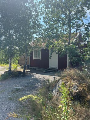 A small, single-story building with a door and window, surrounded by trees and gravel.