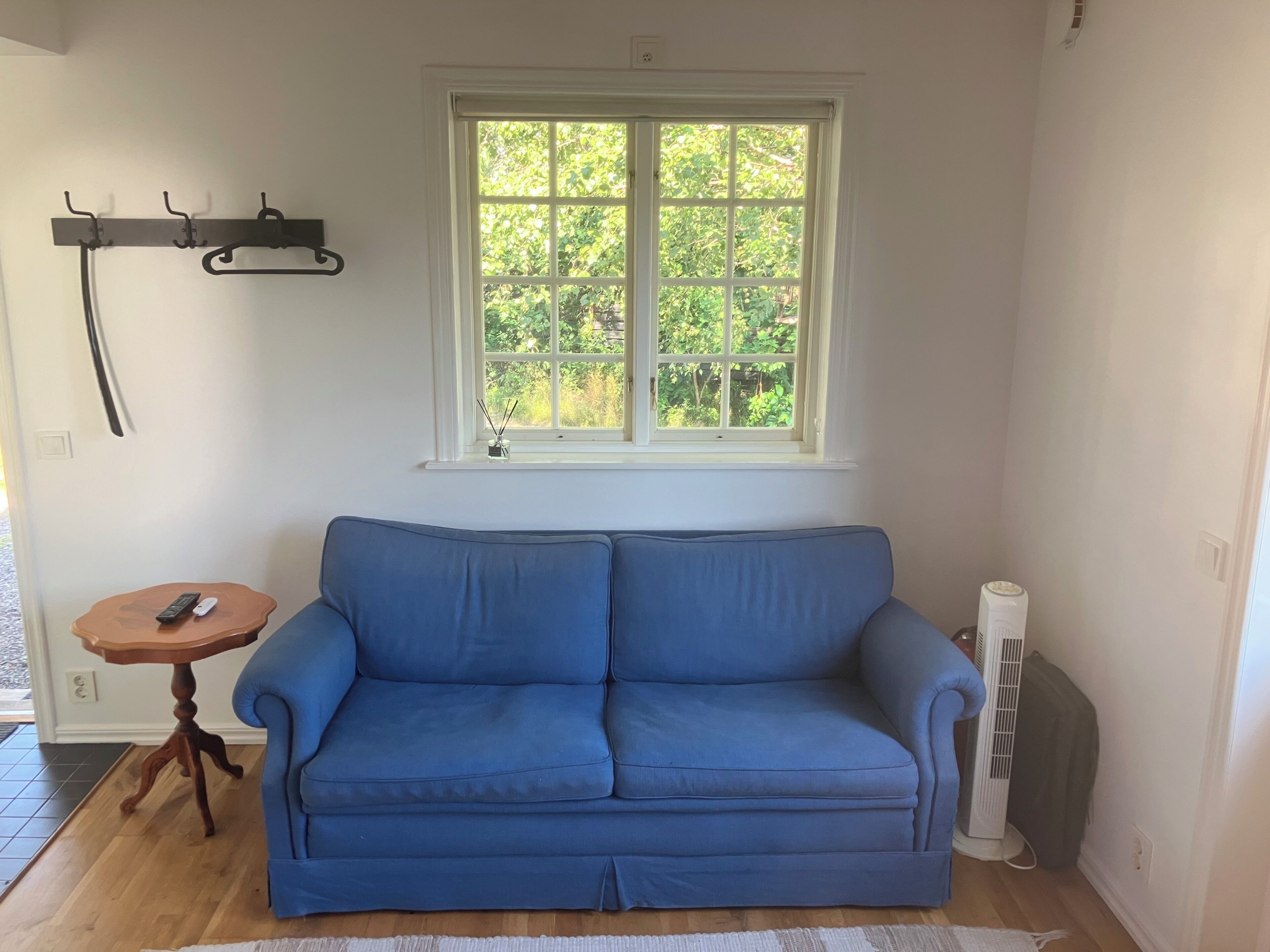 Um quarto com um sofá azul, uma pequena mesa de madeira com um controle remoto, uma janela com vista para o verde e um radiador branco.