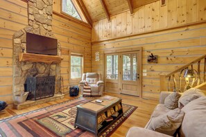 A log cabin interior with a stone fireplace, a sofa, a recliner, and a bookshelf.