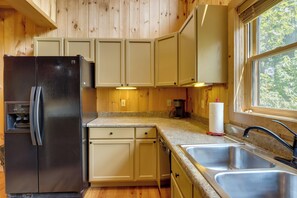 A kitchen with a black refrigerator, beige cabinets, granite countertops, a stainless steel sink, and a window with a view of trees.