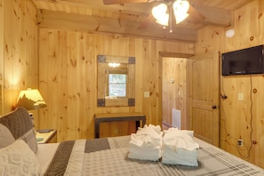 Ein Zimmer mit Holzwänden, einem Bett, einem Schreibtisch, einem Spiegel, einem Fernseher und einem Fenster.