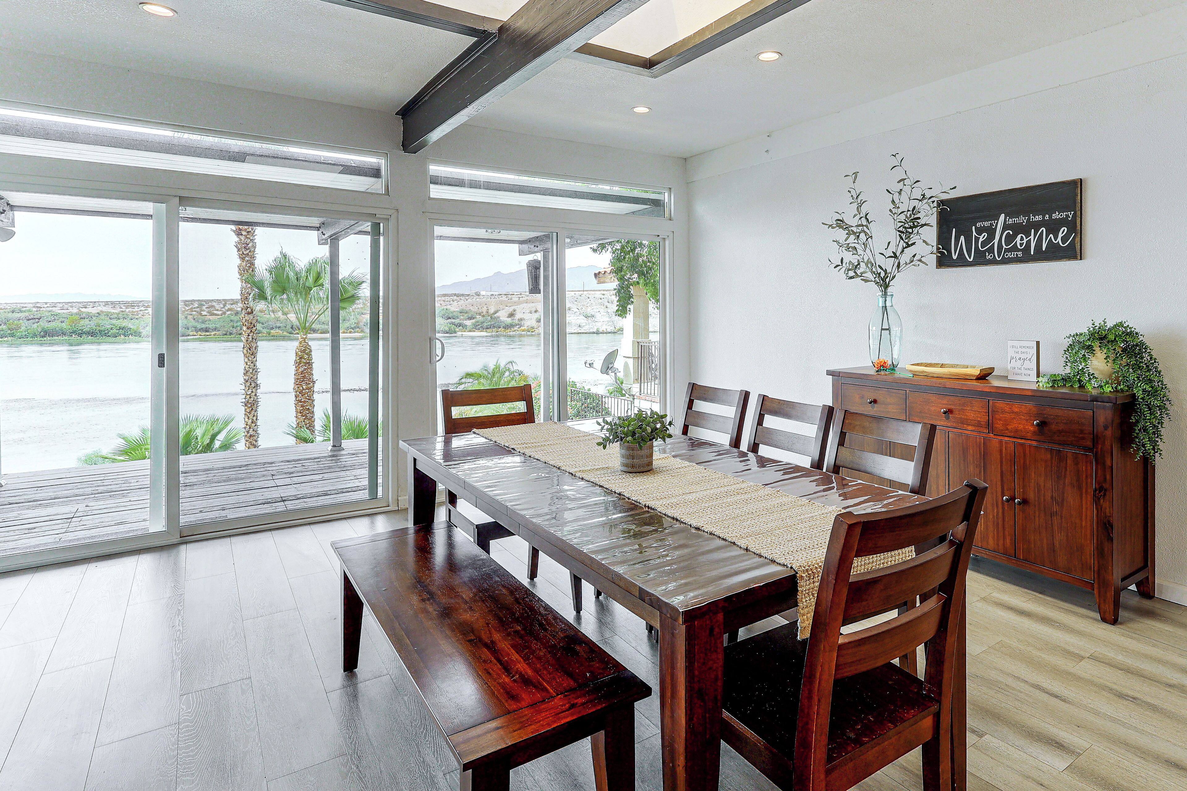 A dining area with a wooden table and chairs, a view of a body of water through large glass doors, and a welcome sign on the wall.