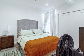 Ein Schlafzimmer mit Bett, Nachttisch, Nachttischlampe, Vorhängen und einem Standventilator.