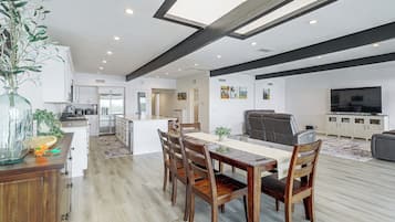 A modern kitchen with white cabinets, a dining area with a wooden table and chairs, and a living room with a sofa and a TV.